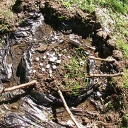 Installation auf einer Wiese. In der Mitte ein Kreis aus Gras darum Wasser auf einer Plane. Mehrere Stöcke verbinden die Mitte mit der restlichen Wiese.