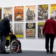 Drei Personen, eine davon im Rollstuhl, stehen vor einer Wand mit politischen Plakaten.
