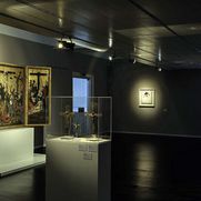 Ausstellungsansicht, im Hintergrund sind drei Kruzifixe und ein mittelalterliches Altarbild zu sehen.