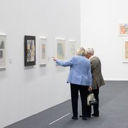 Zwei Personen stehen vor einer Wand mit vier aufgehängten Werken und betrachten diese. Die linke Person zeigt auf eines der Werke und die Personen unterhalten sich.
