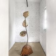 Die Installation von Cynthia Wijono "Nest III" ist zu sehen. Die Installation besteht aus einer Metallstange, auf der drei kugelförmige Gestalten angebracht sind. Am Fuß der Skulptur befindet sich eine größere Kugel. Die Installation ist in den Farben Braun, Weiß, Grün und Goldbraun gehalten. Die Kugeln bestehen aus verschiedenen Materialien wie Karton, Draht, selbstgeschöpftem Papier, Blättern, Rinde, Tannennadeln, Schellack und Müll.