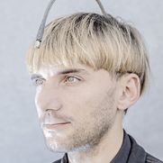 3/4 Portrait eines jungen Mannes mit blonden Haaren. Auf dem Kopf trägt er eine Prothese, die in einem Halbbogen vom Hinterkopf beginnt und über seiner Stirn endet. Sie ermöglicht ihm das farbige Sehen.