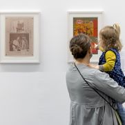 Person mit Kind auf dem Arm steht vor einer Wand mit zwei Werken und betrachtet diese.